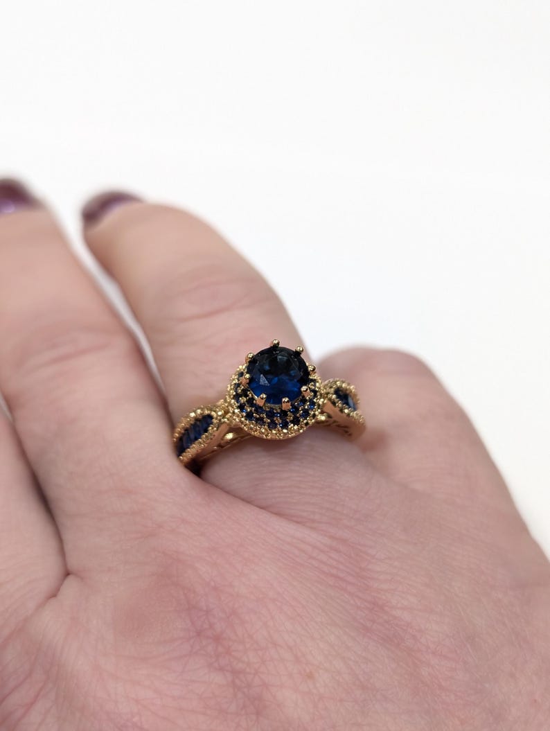 Peut inclure: Une bague en or avec une grande pierre ovale bleue entour&eacute;e de petites pierres bleues. La bague est port&eacute;e sur un doigt.