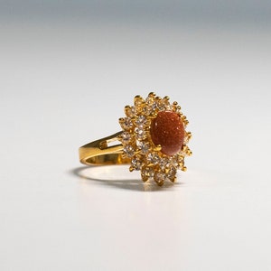 May include: A gold-toned ring featuring a central oval-shaped, reddish-brown stone, surrounded by a halo of smaller, clear gemstones. The ring's band is a simple, polished gold. The design is reminiscent of a flower.