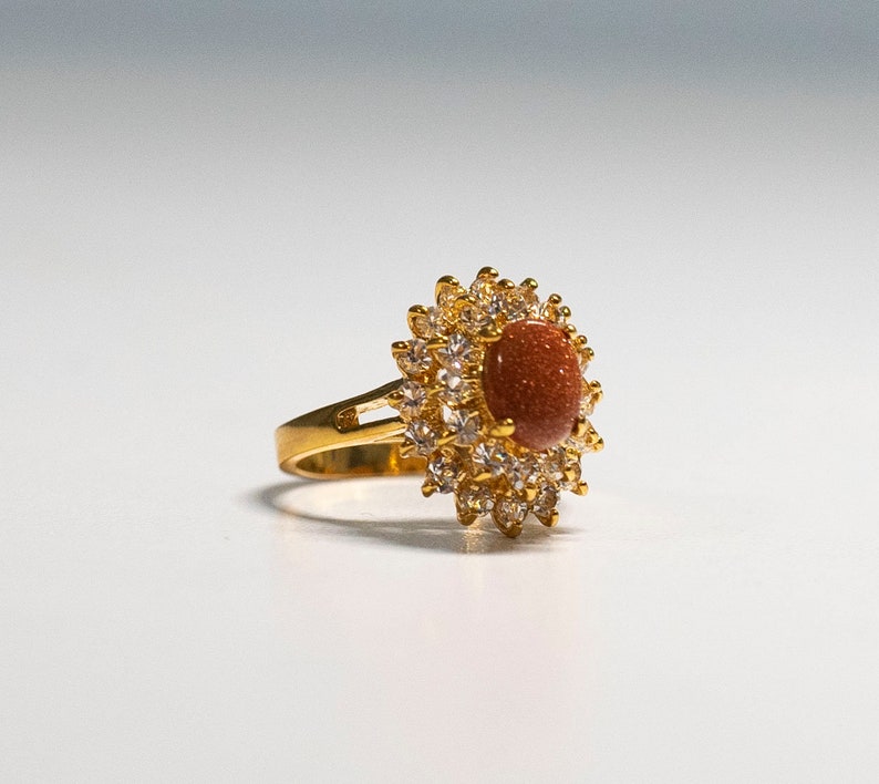 May include: A gold-toned ring featuring a central oval-shaped, reddish-brown stone, surrounded by a halo of smaller, clear gemstones. The ring's band is a simple, polished gold. The design is reminiscent of a flower.