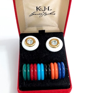 May include: A pair of white circular earrings with gold-tone centers and clear crystals, presented in a red box labeled K.J.L. Kenneth Jay Lane. Below the earrings are ten small, colorful, circular rings in green, red, blue, black, and orange.