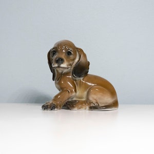 Może przedstawiać: Porcelanowa figurka siedzącego szczeniaka rasy dachshund w kolorze brązowym i białym, z głową przechyloną na bok.