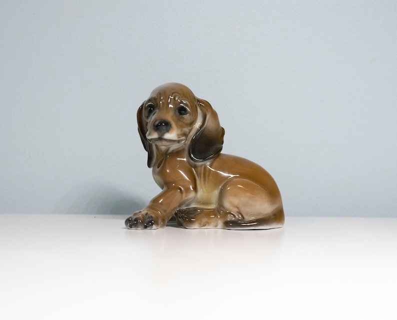 Może przedstawiać: Porcelanowa figurka siedzącego szczeniaka rasy dachshund w kolorze brązowym i białym, z głową przechyloną na bok.
