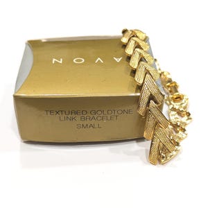 May include: A gold-toned link bracelet with a textured design, next to a gold box with the brand name "AVON" and the text "TEXTURED GOLDTONE LINK BRACELET SMALL". The bracelet has a chevron pattern.
