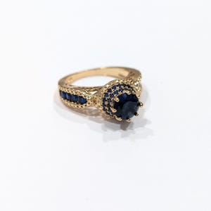 Peut inclure: Une bague en or avec une grande pierre pr&eacute;cieuse bleue entour&eacute;e de petites pierres bleues. La bague a un design d&eacute;licat et complexe.