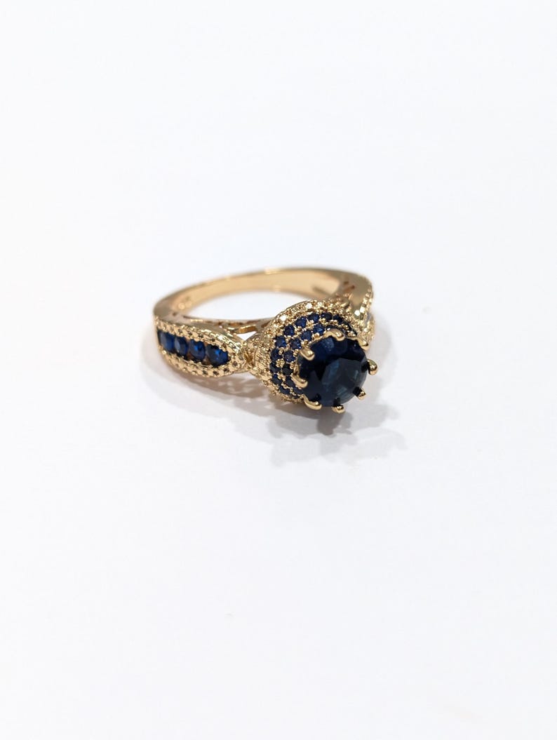 Peut inclure: Une bague en or avec une grande pierre pr&eacute;cieuse bleue entour&eacute;e de petites pierres bleues. La bague a un design d&eacute;licat et complexe.