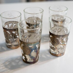 May include: Set of six clear glass shot glasses with silver-toned metal rims and etched designs. The glasses are arranged in a row on a white surface.