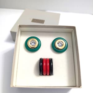May include: A set of two round green earrings with gold-tone centres and clear rhinestones, accompanied by a stack of four rings in alternating black and red colours. The items are presented in a light-coloured box with the Avon logo printed on the lid.