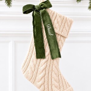 May include: A cream-colored cable knit Christmas stocking featuring a folded cuff and a hanging loop. A dark green velvet ribbon is tied in a bow at the top, with the word Dad embroidered in white thread on one of the ribbon tails.