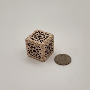 May include: A small, intricately carved wooden cube with openwork geometric designs. The cube is light brown and sits next to a quarter for size comparison. The cube's sides feature repeating circular patterns.