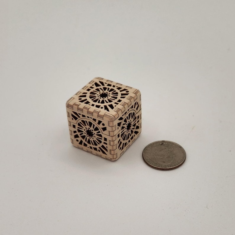 May include: A small, intricately carved wooden cube with openwork geometric designs. The cube is light brown and sits next to a quarter for size comparison. The cube's sides feature repeating circular patterns.