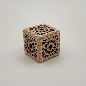 May include: A small, square wooden box with intricate laser-cut geometric patterns. The box is light brown and features detailed designs on all sides, including the top and bottom. The box is likely used for storing small items.