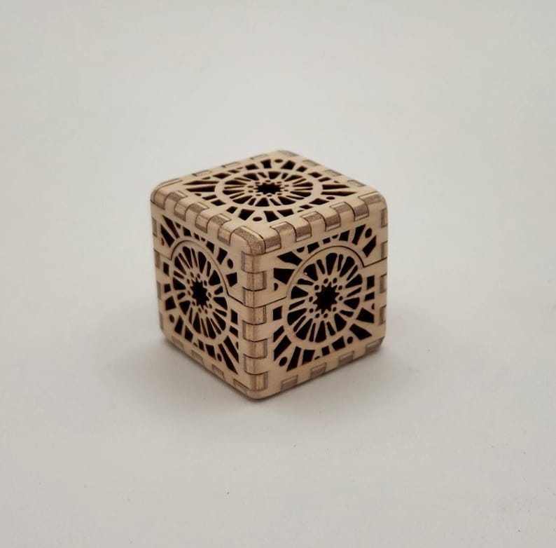 May include: A small, square wooden box with intricate laser-cut geometric patterns. The box is light brown and features detailed designs on all sides, including the top and bottom. The box is likely used for storing small items.