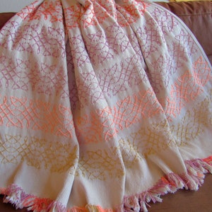 Może przedstawiać: Biały koc z frędzlami. Koc jest wyszywany powtarzającym się wzorem geometrycznych kształtów w kolorze różowym, pomarańczowym i złotym.