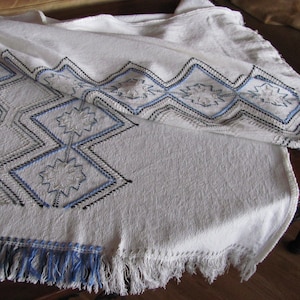 May include: White tablecloth with blue and black embroidered geometric patterns. The tablecloth has a fringed edge.