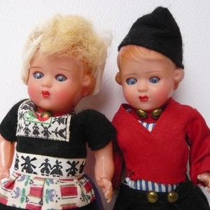 May include: Two vintage dolls dressed in traditional Dutch clothing. The girl doll has blonde hair and wears a black dress with a white and red skirt. The boy doll has brown hair and wears a red jacket with a blue and white striped shirt and black pants.