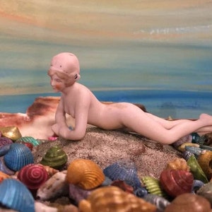 Puede incluir: Una figurita de porcelana de una mujer acostada sobre una cama de conchas de mar de colores. La mujer lleva un traje de baño rosa y tiene una gorra blanca en la cabeza.