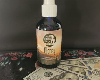 May include: A dark blue glass spray bottle with a white top, labeled "Wolf Spirit Sage Room Spray - Money." The label features a landscape design. Several $100 bills are visible in the foreground.