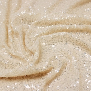 Puede incluir: Primer plano de una tela color crema cubierta de pequeñas lentejuelas redondas. La tela está drapeada, creando pliegues y sombras suaves. Las lentejuelas captan la luz, dando a la tela un aspecto brillante. Ideal para ropa o decoración de eventos.