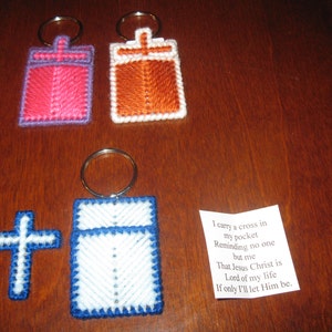 May include: Three keychains with cross designs. The keychains are made of plastic canvas and feature a cross design with a white background and blue, pink, and orange accents. The keychains are hanging on a wooden surface. A small white cross and a note with a religious message are also visible.