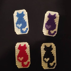 Puede incluir: Cuatro pequeños paneles rectangulares de lienzo blanco con un diseño de punto de cruz de una silueta de gato en diferentes colores. Los gatos son azul, morado, rojo y negro.