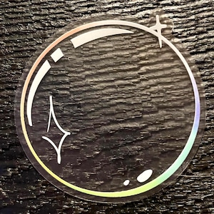 May include: Clear sticker with a circular design. The sticker features a white outline of a bubble with a star and small dots. The edges of the sticker have a rainbow iridescent effect.