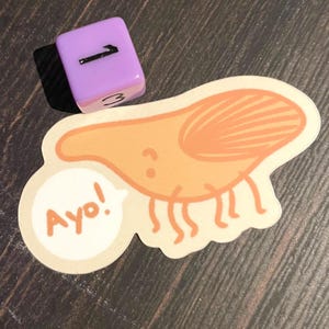May include: A sticker with a cartoon shrimp illustration in orange and white, with the word "Ayo!" in a speech bubble. A purple die with the number 1 visible is next to the sticker.