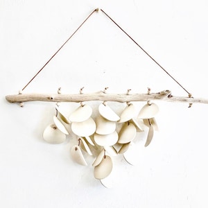 May include: A decorative wall hanging featuring a natural driftwood branch with small wooden beads. Cream-colored, leaf-shaped elements are suspended from the branch by thin cords, creating a layered, cascading design. The piece is suspended by a brown cord.
