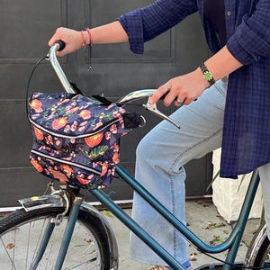 May include: A dark blue bicycle with a floral patterned handlebar bag featuring orange and pink flowers on a dark background. The bag has two zippered compartments and is attached to the silver handlebars of the teal bicycle frame.