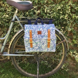 May include: A blue and white floral patterned backpack with brown leather straps and buckles attached to a bicycle rack.