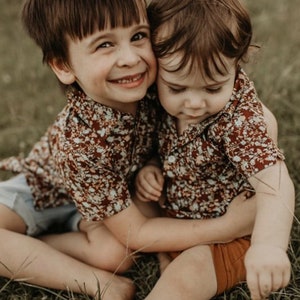 May include: Two children wearing matching floral print shirts in red, brown, and cream tones. They are sitting on grass, embracing. One child smiles, the other looks down. The shirts have short sleeves.