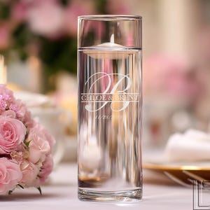 May include: A clear glass cylinder vase filled with water, holding a floating candle. The vase is etched with the names "Chloe & Brent" and a date. The background features a soft focus of pink flowers, suggesting a wedding or event setting.