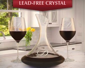 May include: A set of two clear glass wine glasses and a crystal decanter, all engraved with the letter "D". The glasses are filled with red wine and the decanter is filled with red wine. The decanter has a wide base and a narrow neck. The glasses have a long stem and a round bowl. The set is on a white countertop.