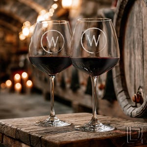 May include: Two clear glass wine glasses filled with deep red wine, each featuring a white circular emblem with the letter W etched on the bowl. The glasses have long, slender stems and sit on a rustic wooden surface in a dimly lit environment.