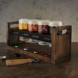 May include: A wooden beer flight rack with five glasses, a bottle opener, and six bottle caps. The glasses are filled with different colored beers and labeled with the names "IPA", "Lager", "Amber", and "Stout". The rack is personalized with the name "Roberts".