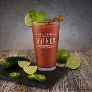 May include: A personalized glass with the name "WALKER" etched on it. The glass is filled with a red beverage and garnished with green olives and celery. The glass is sitting on a black slate surface with green jalapeno peppers and lime slices.