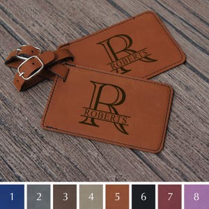 May include: Two brown leather luggage tags with a black leather strap. The tags are personalized with the letter "R" and the name "ROBERTS" in a script font. The tags are shown against a wood background. There are eight color swatches below the tags.