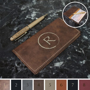 May include: A brown leather checkbook cover with a stitched edge and a monogrammed initial "R" in a circle. The cover is lying on a black and white marble surface. A wooden pen with the name "ROBERTSON" engraved on it is lying next to the cover.