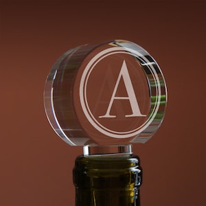 May include: A clear glass bottle stopper with a circular design featuring the letter "A" in a circle. The stopper is on top of a wine bottle.