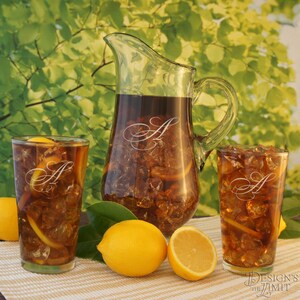 May include: A glass pitcher and two glasses filled with iced tea. The pitcher and glasses are etched with the letter "A". There are lemons on the table next to the glasses.