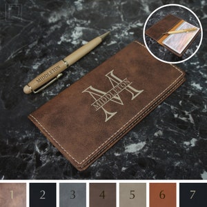 May include: A brown leather checkbook cover with a personalized monogram and the name "Middleton" engraved on it. The cover has a stitched edge and is shown with a wooden pen.