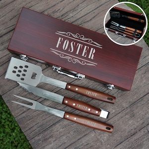 May include: A wooden box with a personalized engraved name "FOSTER" on the lid. The box contains a set of four grilling tools with wooden handles, including a spatula, a fork, and two tongs.