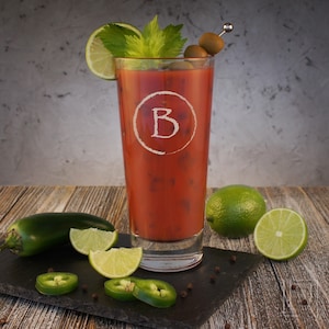 May include: A tall glass filled with a red bloody mary cocktail garnished with celery, olives, and lime wedges. The glass has a white circle with the letter "B" in the center.