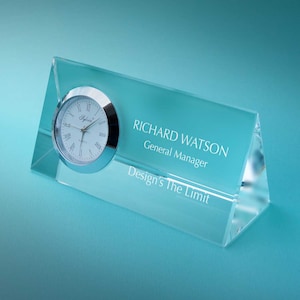 May include: A clear glass desk clock with a silver face and hands. The clock is engraved with the text "RICHARD WATSON, General Manager, Design's The Limit".