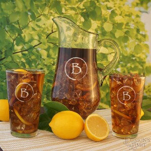 May include: A glass pitcher and two glasses filled with iced tea. The pitcher and glasses are etched with the letter "B" inside a circle. There are lemons on the table next to the glasses.