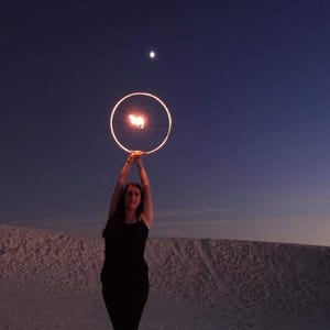May include: A person in a black dress is holding a glowing hoop in the air. The hoop is lit with fire and is in front of a dark blue sky with a small moon in the distance.