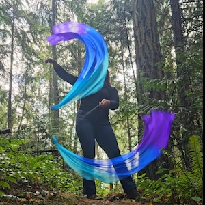 May include: A person is holding two flowing silk flags in a forest setting. The flags are a gradient of blue and purple. The person is wearing a black long-sleeve shirt and dark blue pants.