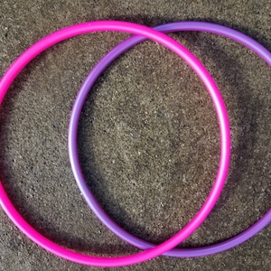 May include: Two plastic hula hoops, one pink and one purple, are intertwined on a gray concrete surface.