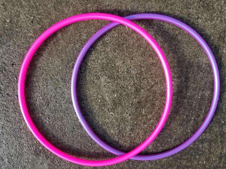 May include: Two plastic hula hoops, one pink and one purple, are intertwined on a gray concrete surface.