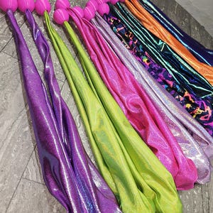 Könnte beinhalten: Eine Sammlung farbenfroher, schimmernder Tanzbänder mit rosa Kugelgriffen. Die Bänder sind in verschiedenen Farbtönen erhältlich, darunter Lila, Grün, Pink und Orange. Der Stoff hat einen metallischen Glanz.