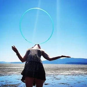 May include: A person is spinning a blue hula hoop above their head while standing on a beach. The person is wearing a black and white patterned dress. The background is a blue sky and a body of water.